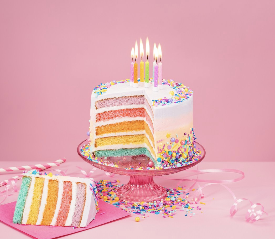 Birthday cake and slice with pastel-colored layers, white icing, and candles against a pink background