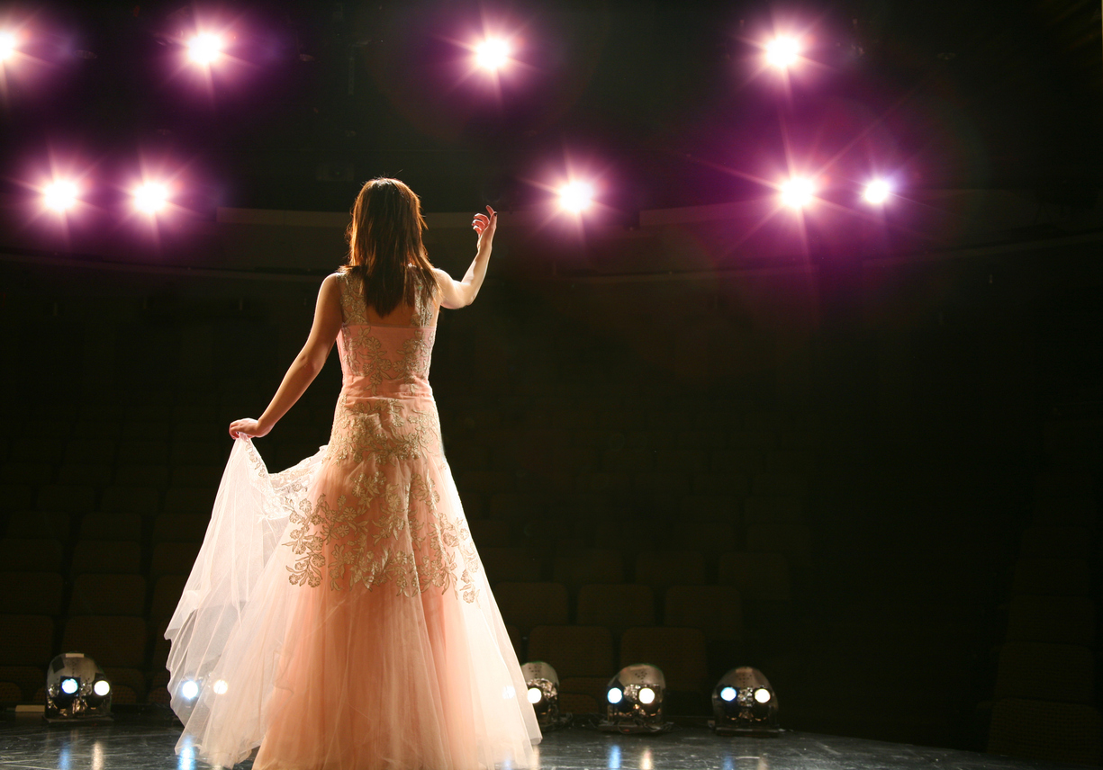 Rear view of adult female singer in formal gown on stage facing spotlights looking into the theater